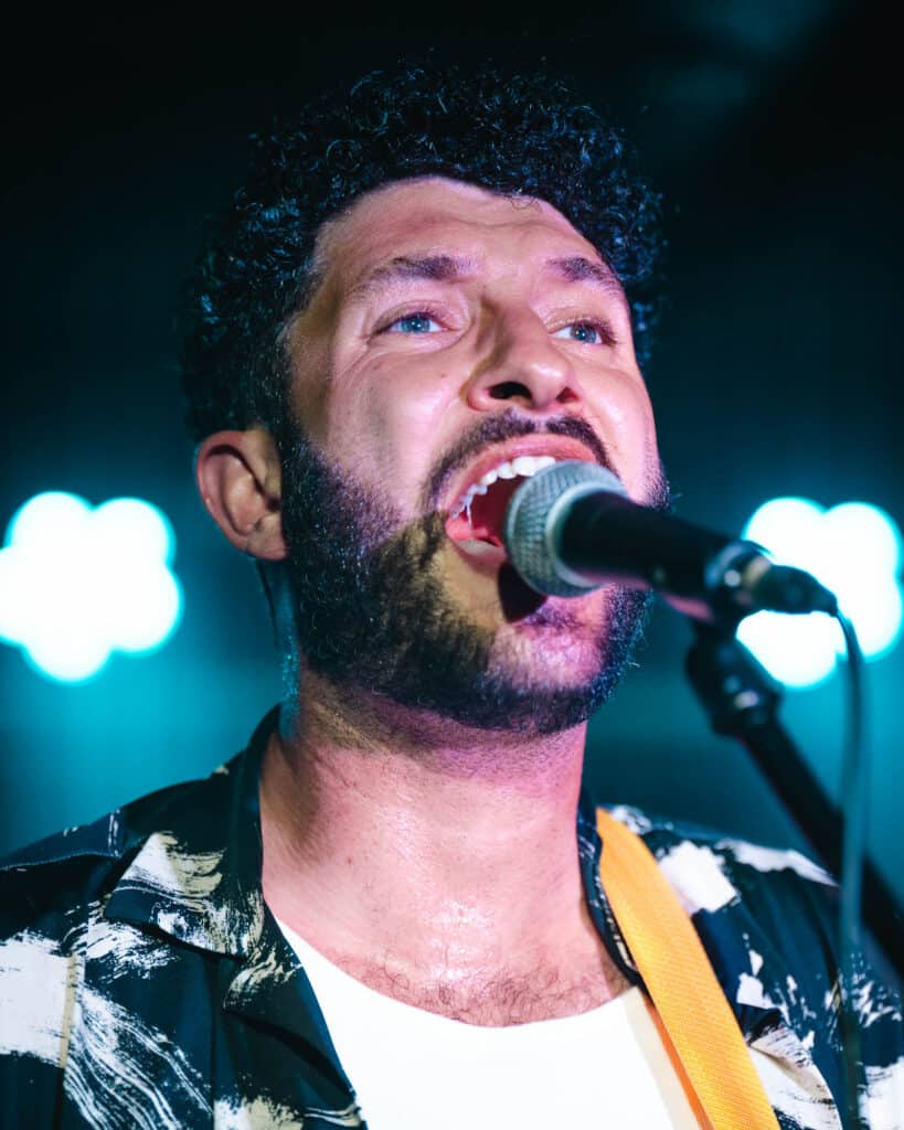 A man with curly hair and a beard sings passionately into a microphone, captured in stunning headshot photography. He wears a patterned shirt, and the background glows with mesmerizing blue lights.