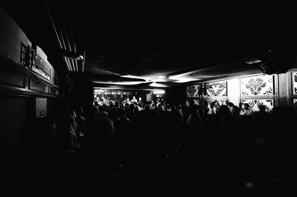 A black and white photo by a professional event photographer captures a crowded music venue, where people are silhouetted against the stages bright lights. The ornate patterned walls to the right add to the lively and energetic atmosphere.