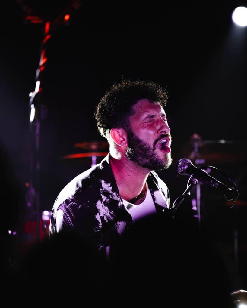 A singer passionately performing on stage in low light, a microphone in front of him. Clad in a patterned shirt, he emits an intense vocal expression, reminiscent of a dynamic professional headshot. Drums form the blurred backdrop to his captivating presence.
