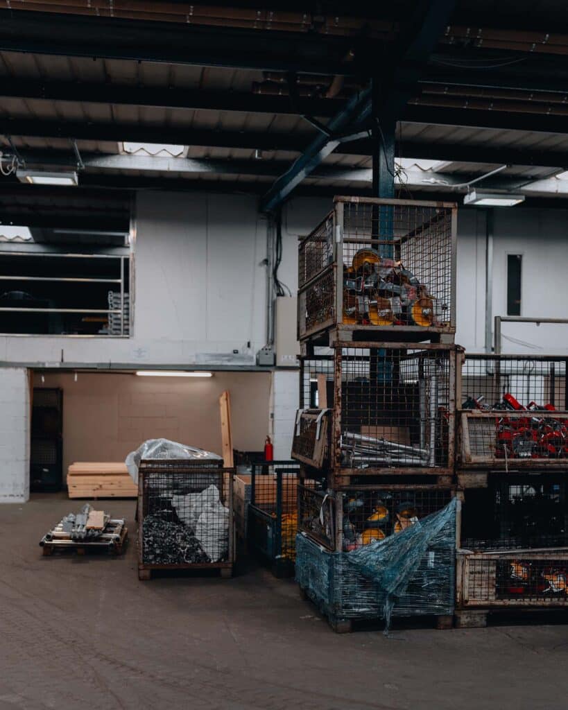 Industrial interior captured with a keen eye for industry photography: metal cages stacked and filled with various tools and machinery parts, while a pallet with materials looms in the background. The spaces high ceiling and large windows enhance its rugged, industrial atmosphere.