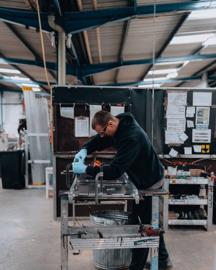 In this industrial photography scene, a person wearing protective gloves diligently works on a piece of equipment in the workshop. Surrounded by tools and materials, with shelves and notices in the background, the metal elements emphasize the settings gritty aesthetic.
