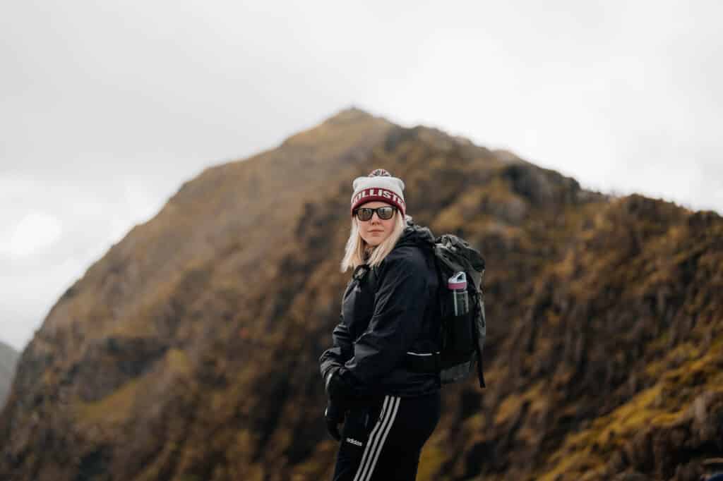 A person in a beanie, sunglasses, and black outdoor attire strikes a pose on a mountain slope with a backpack. The portrait photography captures the misty, rugged landscape under a cloudy sky, highlighting the adventurers spirit amidst natures grandeur.