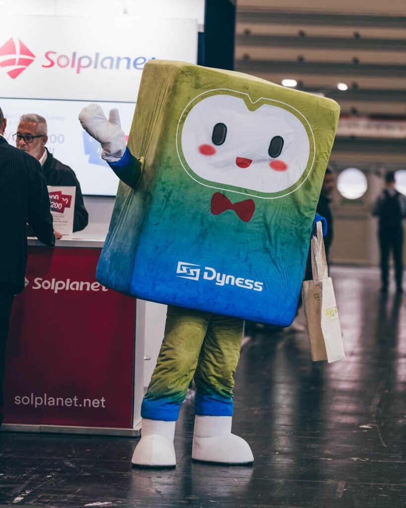 A person in a colorful costume resembling a rectangular character with a smiling face waves and holds a tote bag, perfect for commercial photography. The costume reads Dyness, while in the background, theres a booth adorned with Solplanet signage.
