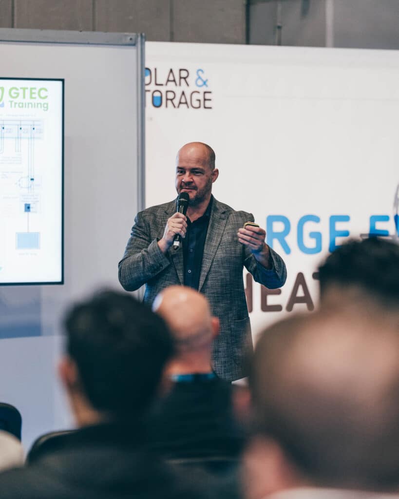 A person, clad in a gray blazer, speaks into a microphone at a conference or seminar. In the background, the screen displays GTEC Training as the blurred audience focuses on this moment of commercial photography.