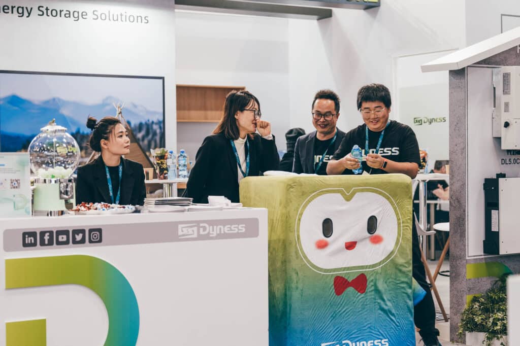Four people at a booth for Dyness Energy Storage Solutions, with two wearing glasses. Their smiles are captured perfectly by professional photography near me. A large green and white mascot with a smiling face stands beside them, while the table is filled with snacks and promotional materials.