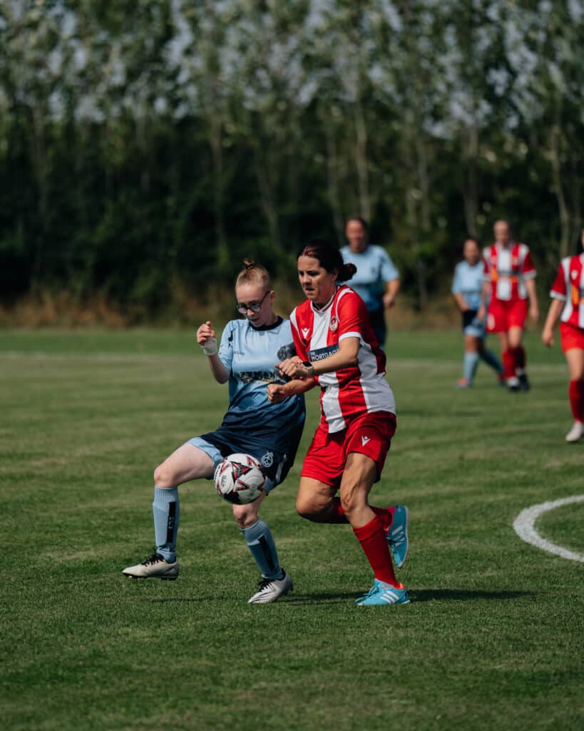 Two soccer players, one in light blue and the other in red and white, fiercely compete for the ball on a grassy field. Positioned for a perfect shoot sports, other players are visible in the background, with trees lining the perimeter.