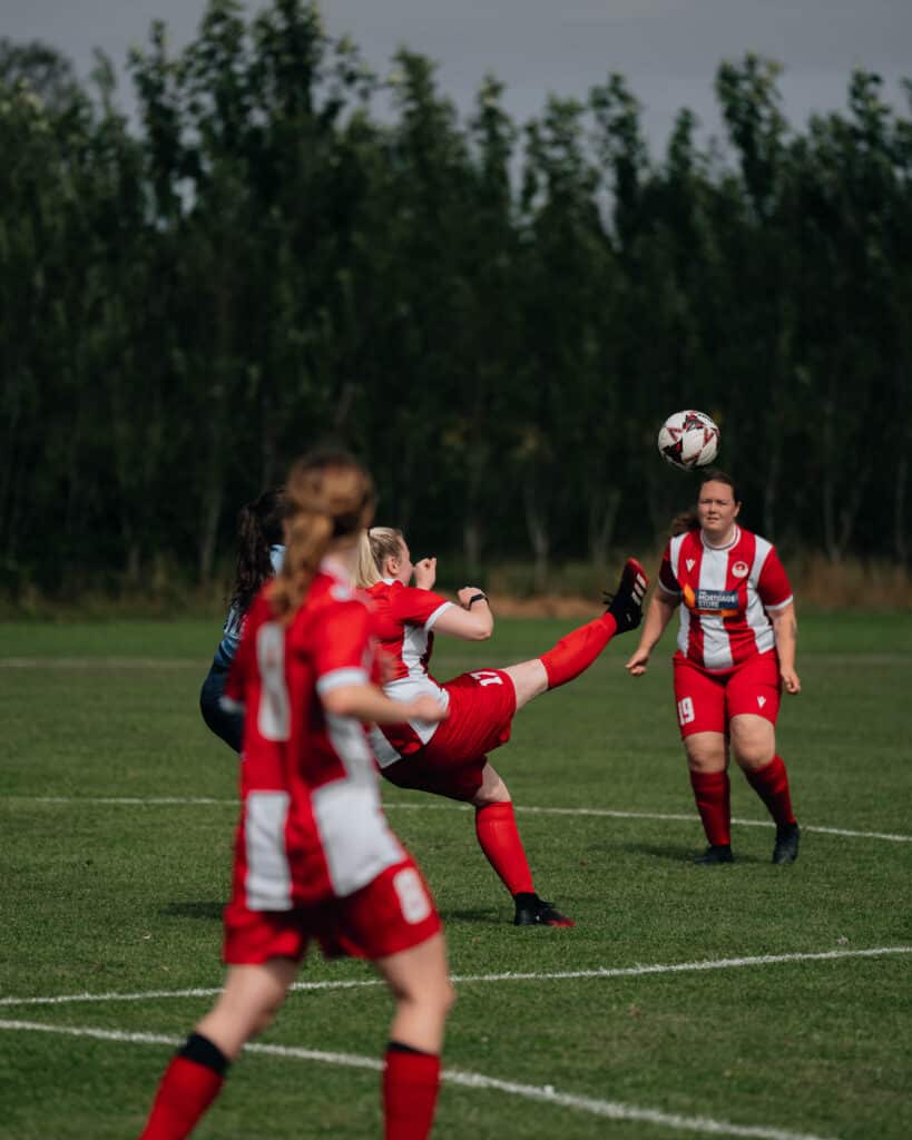 A womens soccer game is captured in stunning sports photography. On a grassy field, a player in red and white kicks the ball mid-air as her teammates and an opponent watch intently. Trees stand tall in the background under a cloudy sky.