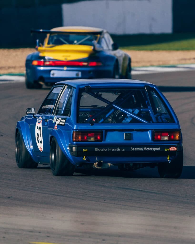 Amidst the thrilling car race, a blue hatchback with the number 63 and a distinct roll cage leads, closely pursued by a yellow sports car. Under the bright sun, both cars gracefully navigate a curve, capturing the essence of high-speed car race photography.